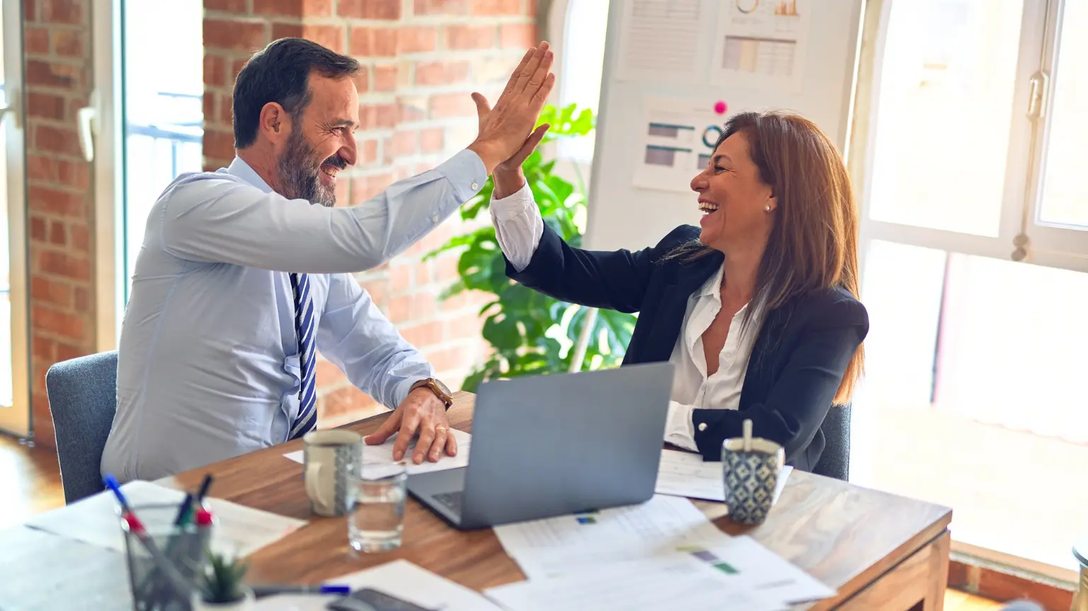 Two coworkers smiling and giving a high-five across a desk with laptops and papers.